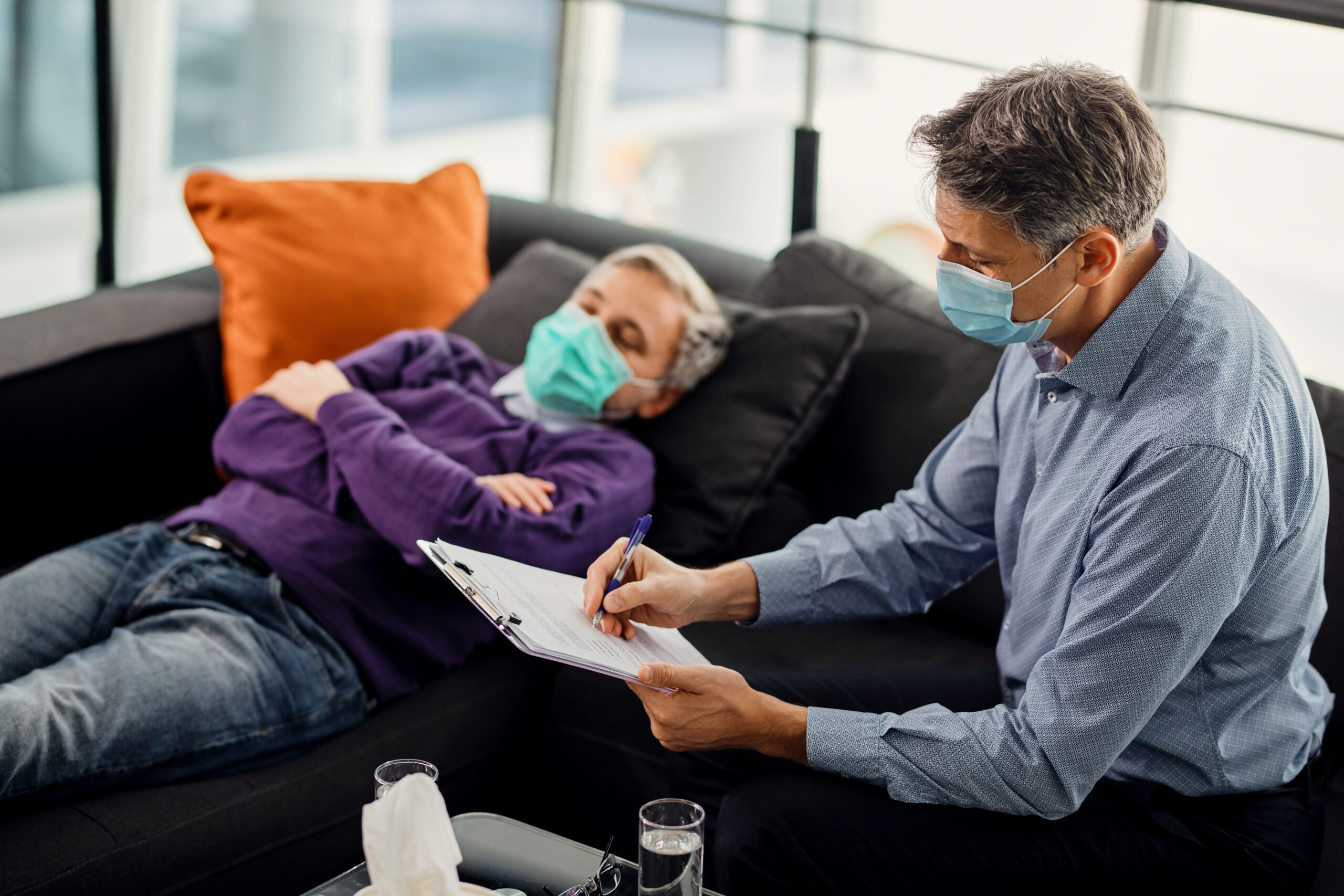 Mental health professional taking notes during a session with his patient. They are wearing protective face masks due to coronavirus pandemic.